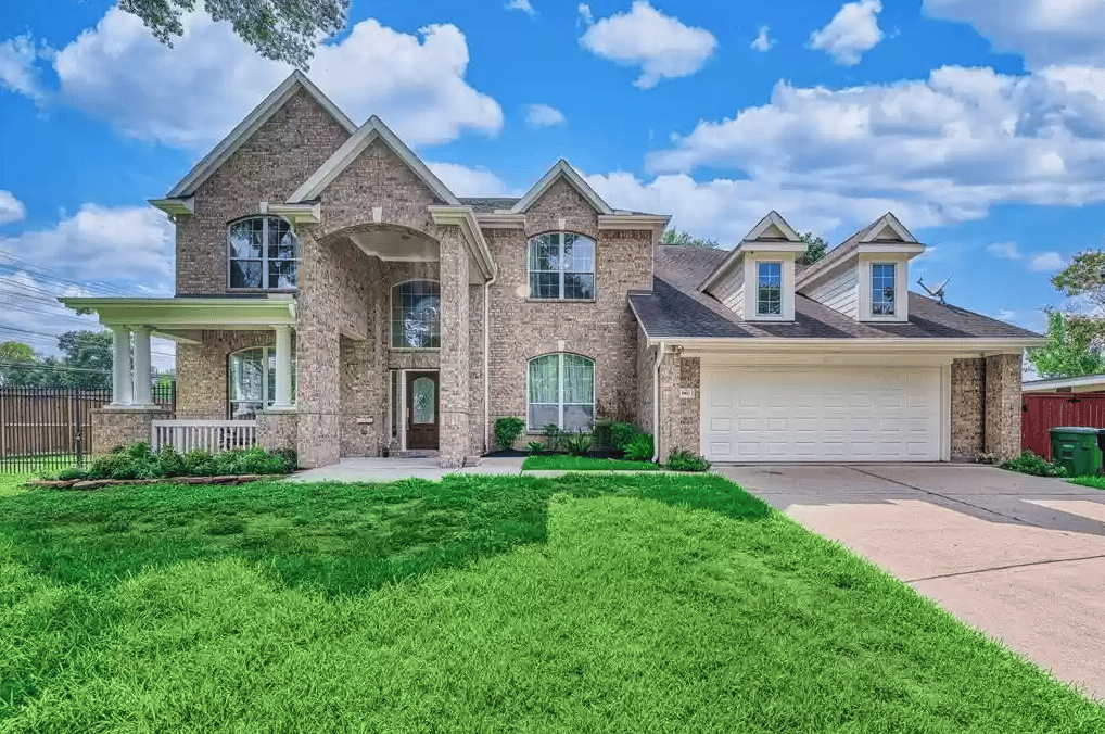 Two-story brick house with a covered entry, white double garage, dormers, and broad green lawn.