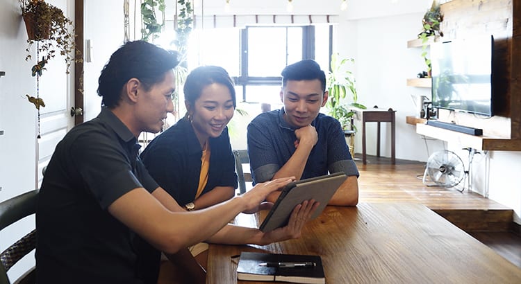 Three people sitting at a table, engaged in a discussion while looking at a laptop. The setting appears to be informal, possibly a meeting or consultation.