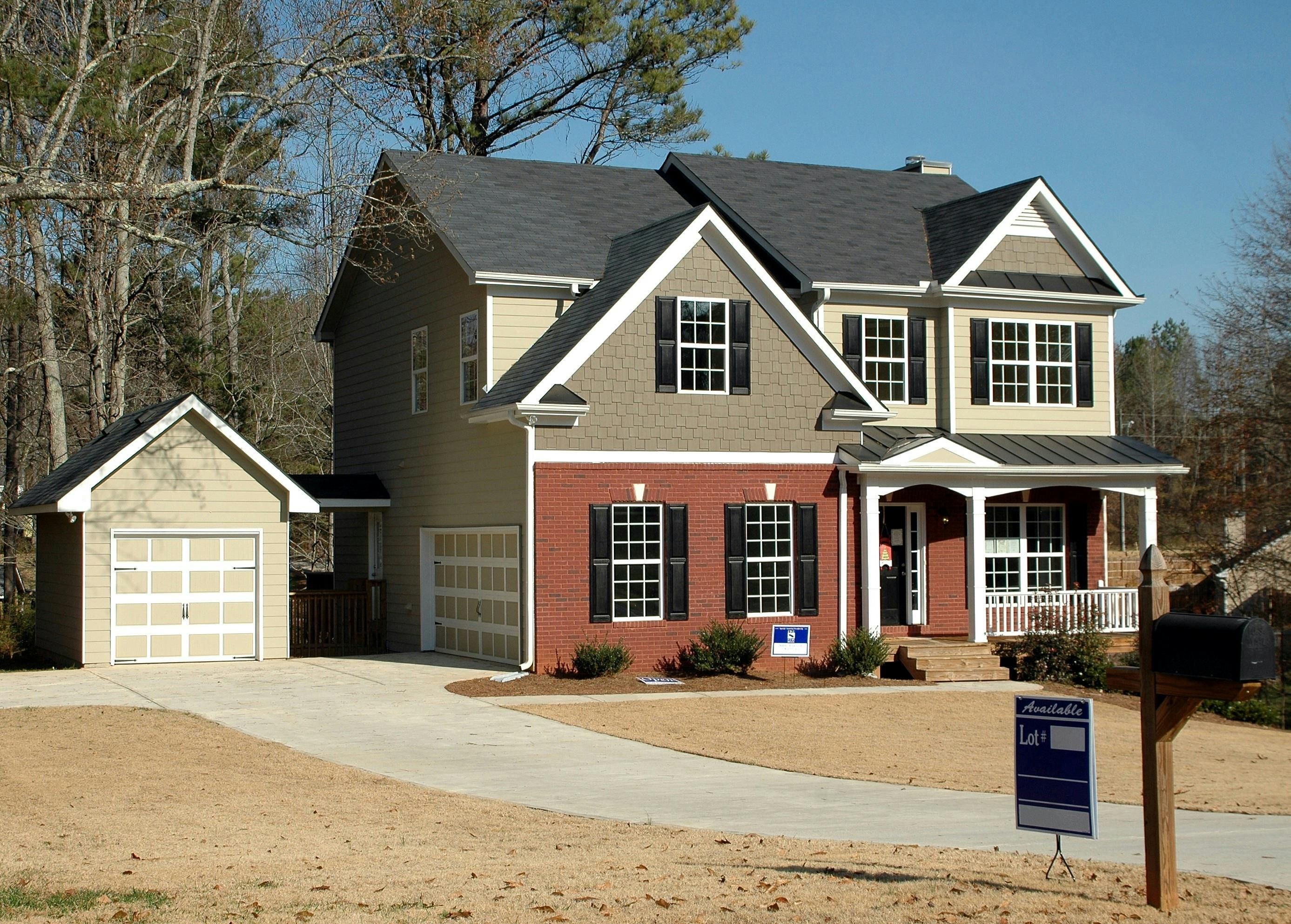 Two-story beige and brick house with an attached garage, covered porch, and available-lot sign.