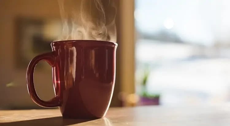 A close-up of a steaming red coffee mug placed on a table. The background is softly blurred, highlighting the warmth and comfort associated with a hot beverage.