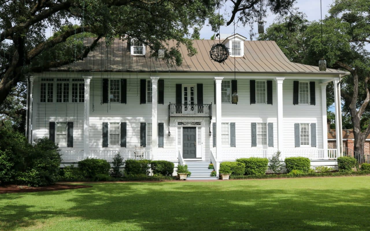 A large white house with black shutters sits on a lush green lawn. The sky is a clear blue, and the sun is shining.