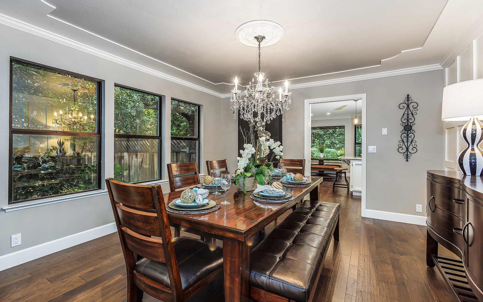 Dining room with a set wood table, crystal chandelier, leather bench, and windows overlooking greenery.