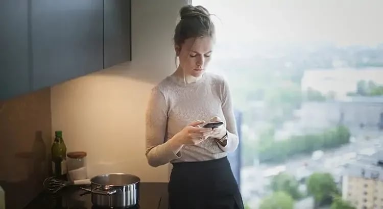 A woman in a kitchen looking at her phone. She appears to be preparing food or a drink, with kitchen items visible around her.