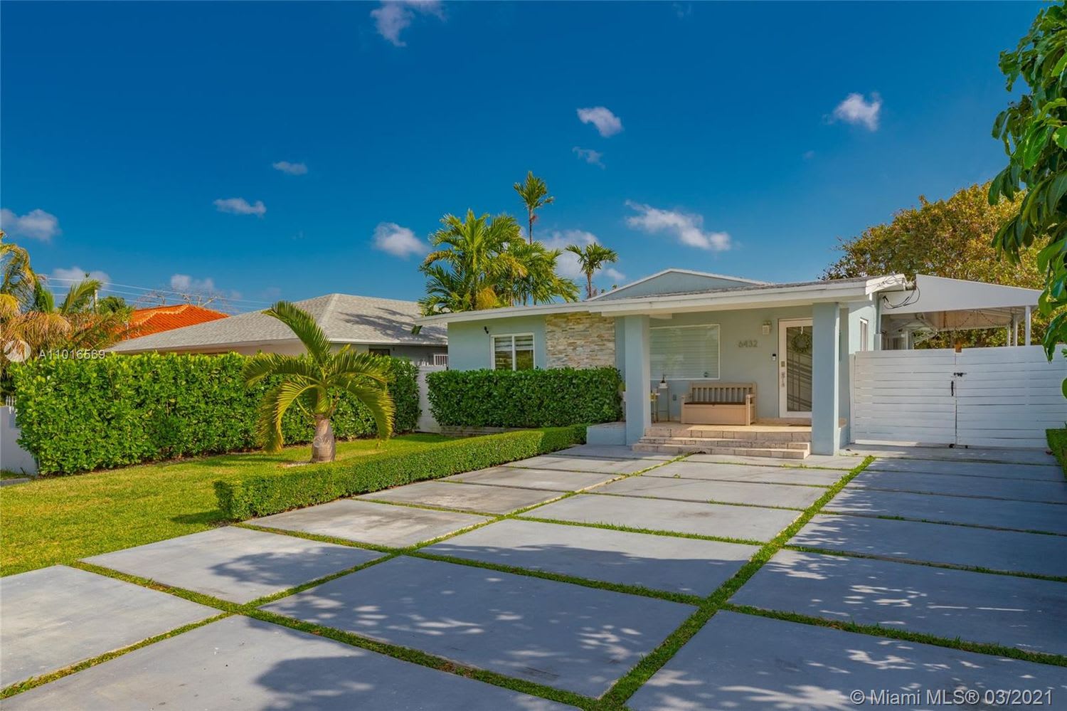 Pale blue single-story home with a stone accent facade, broad driveway, white gate, and manicured tropical landscaping.