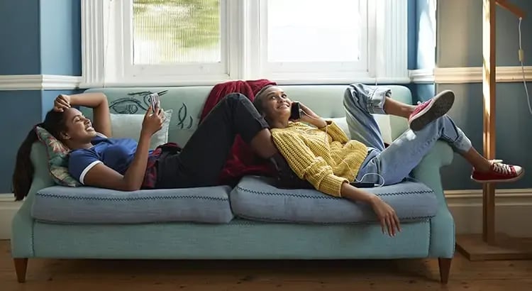 Two people are lounging on a couch, using their phones. One person is lying down with their legs over the arm of the couch, while the other is sitting and leaning back.