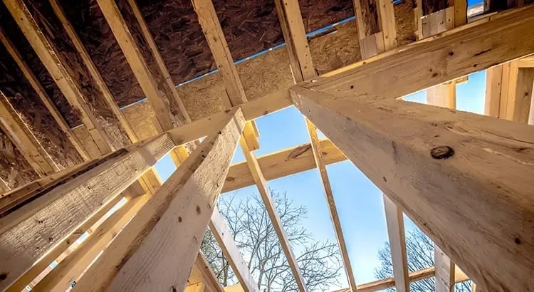 A view of a wooden house frame under construction, with blue sky visible through the beams. This image highlights the process of building a home.