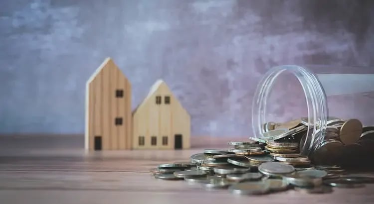 Small wooden house models and stacks of coins next to a glass jar, suggesting saving for a home or financial planning.