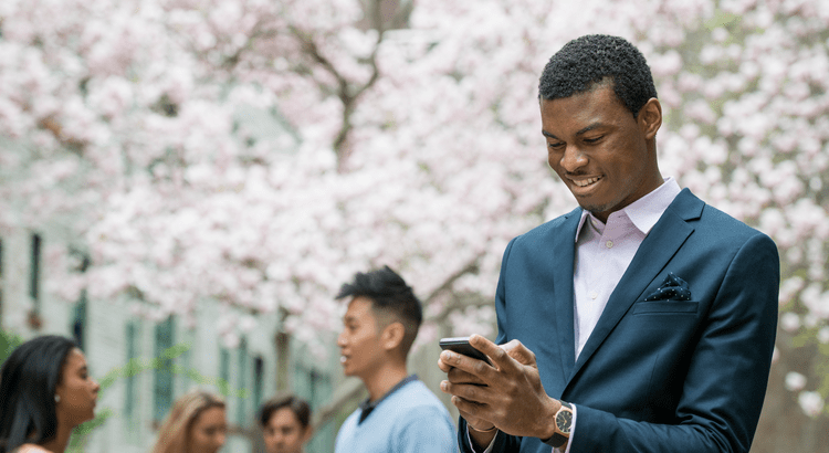 A photo of a man in a suit using a smartphone, with people and cherry blossoms in the background.