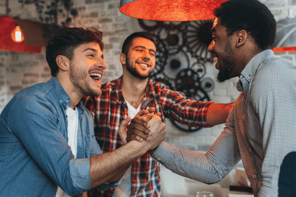 Three men laugh and clasp hands in a friendly greeting beneath a red pendant light.