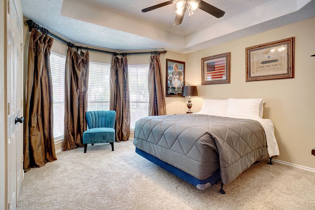 Bedroom with a gray-quilted bed, teal chair, and bay windows framed by brown curtains.