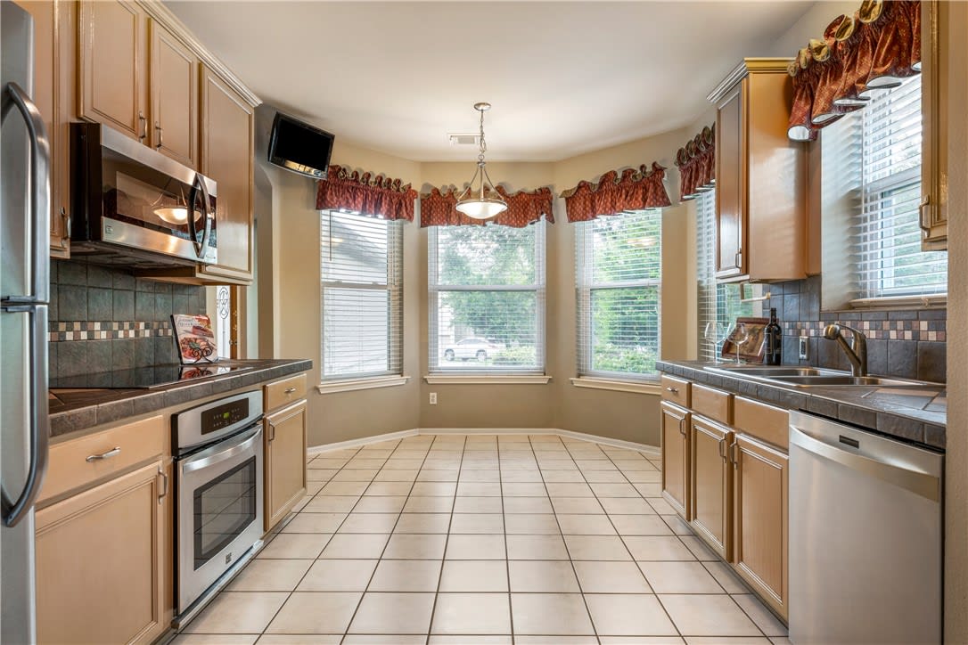 Kitchen with tan cabinetry, stainless steel appliances, and a three-window bay with red valances.