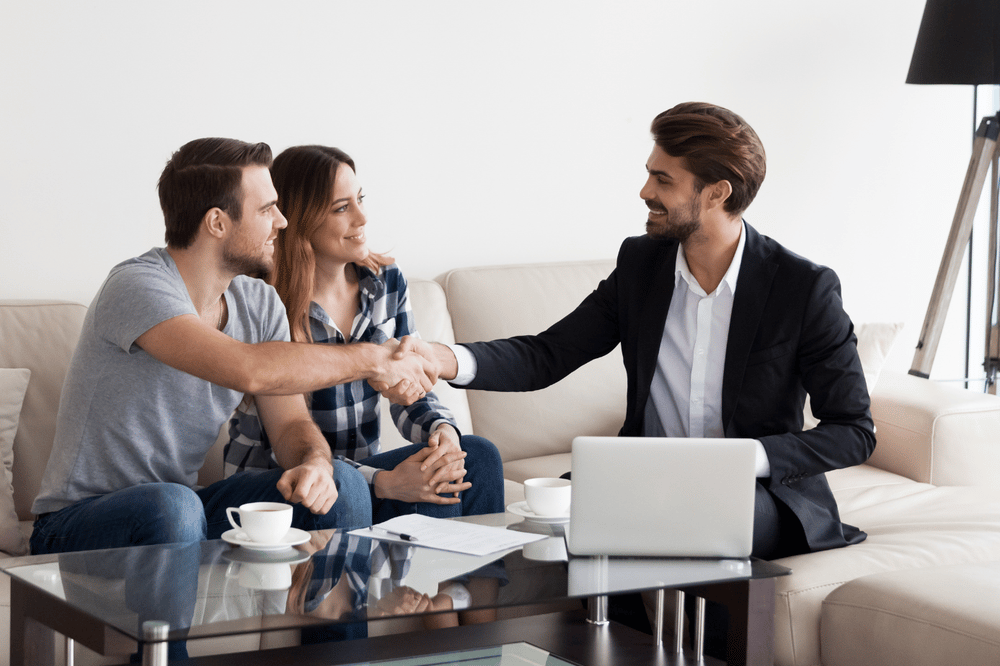 Real estate agent shakes hands with a couple seated on a cream sofa.