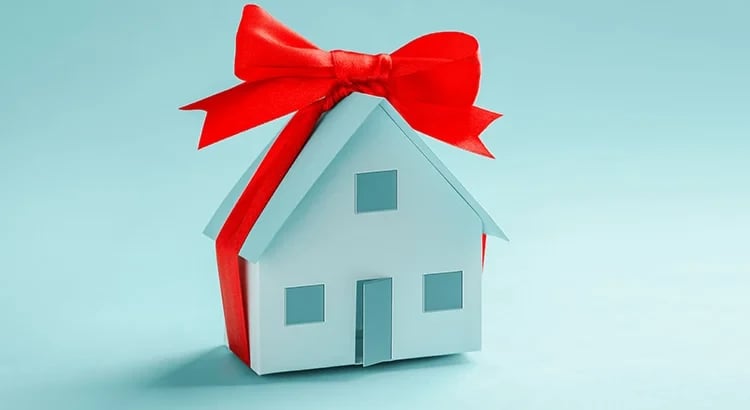 This image features a small model house wrapped with a large red bow, indicating a gift or purchase. The background is blue, and the house is the central focus.