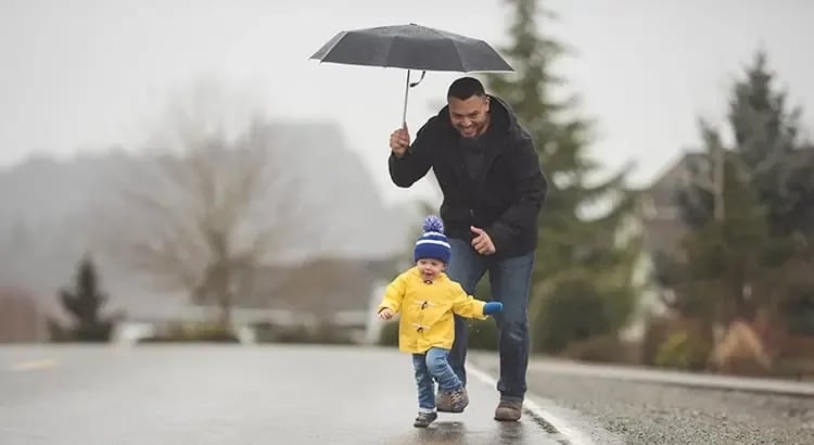 A man holding an umbrella, walking with a child in a yellow raincoat. They appear to be enjoying a rainy day together.