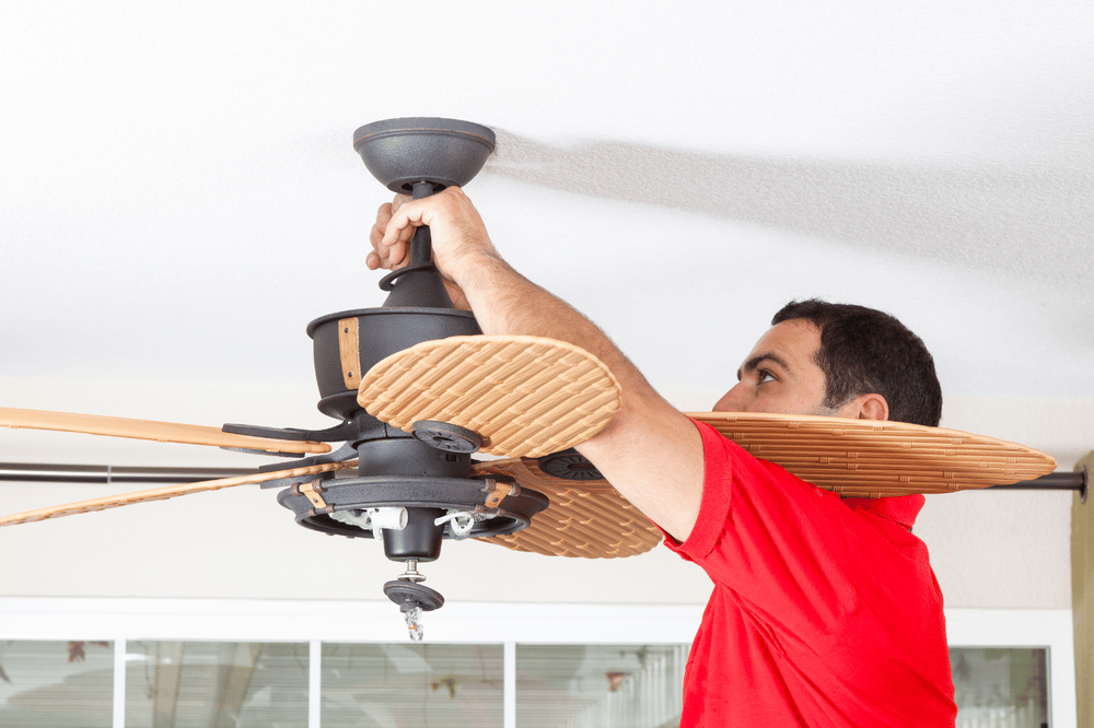 A man installs a ceiling fan, holding its blades beneath the ceiling.