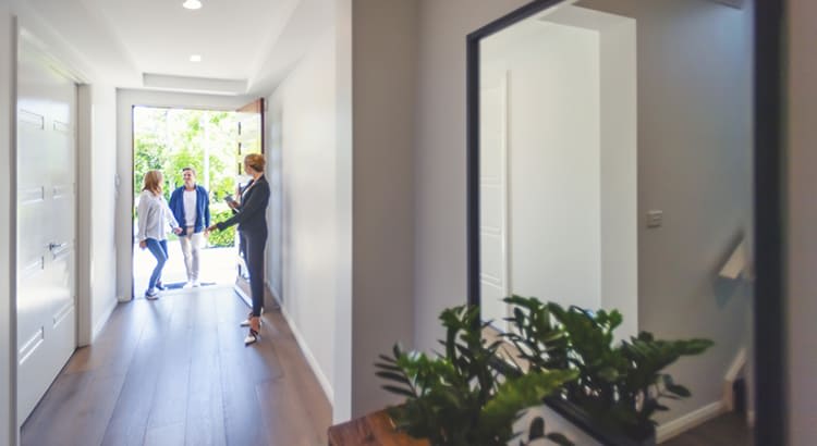 a modern home, specifically a hallway that leads to an open front door where two people are standing, likely guests or potential homeowners. 