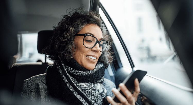 A smiling woman wearing glasses and a scarf, sitting in a car and using a smartphone. 