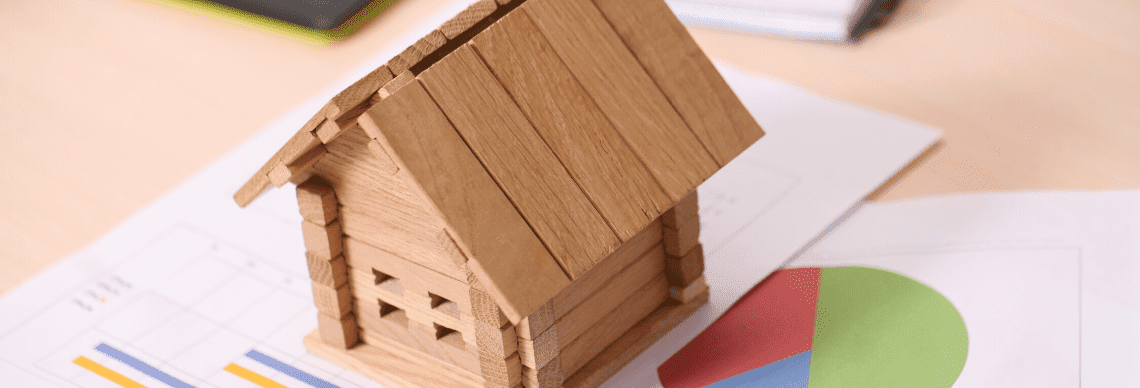 A wooden house model placed on top of documents with charts, suggesting a connection between real estate and financial planning. 