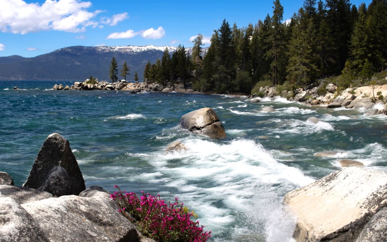 Dramatic Lake Tahoe shoreline with crashing waves, rocky terrain, and snow-capped mountains.