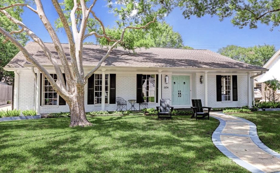 A single-story white house with black shutters, a blue front door, a maintained lawn, trees,  and a paved walkway.