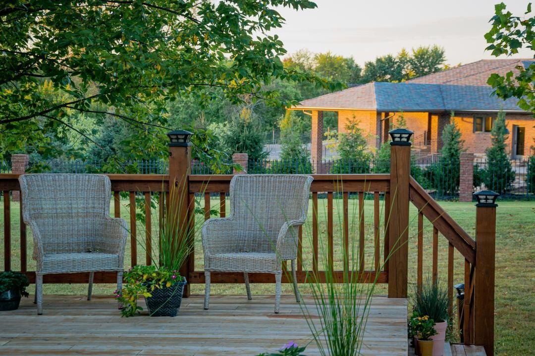 Backyard deck with wicker chairs and potted plants overlooking a lawn and brick house.