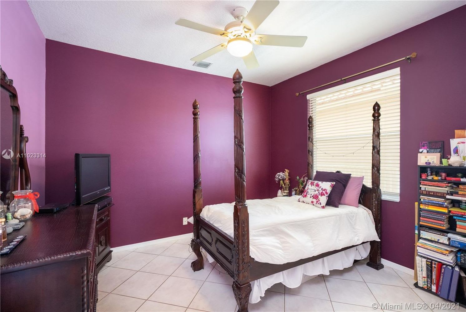 Bedroom with a dark carved four-poster bed, deep purple walls, tiled floors, and a ceiling fan.