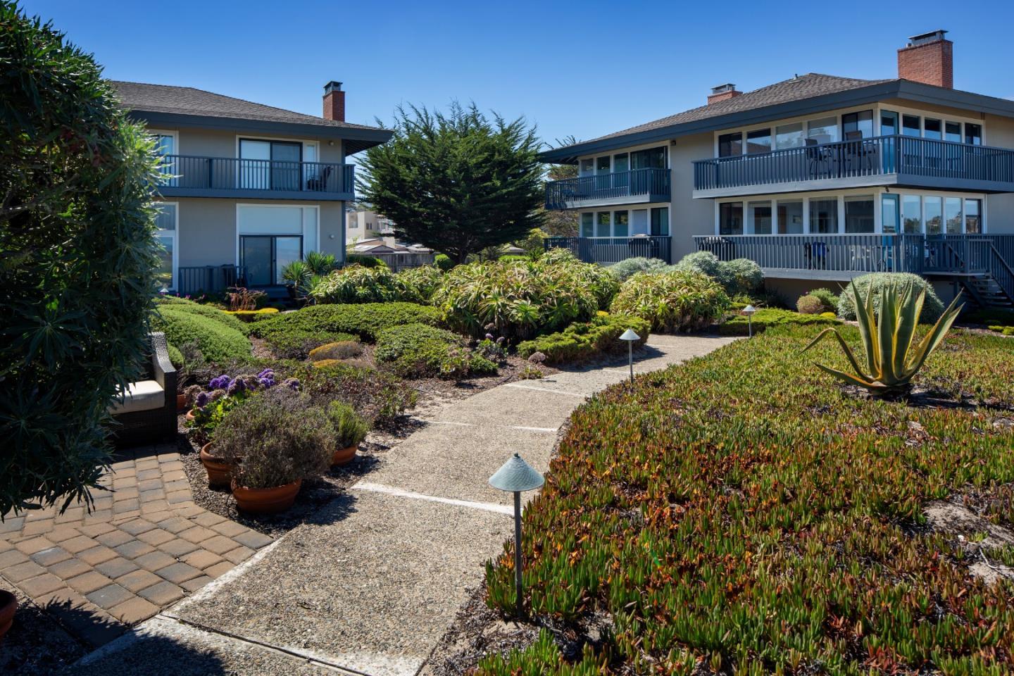 Two gray residential buildings with dark balconies face a landscaped courtyard and paved walkway.