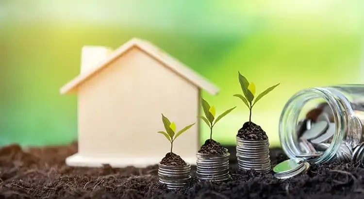 A small house model with growing plants and stacked coins, symbolizing investment and growth.