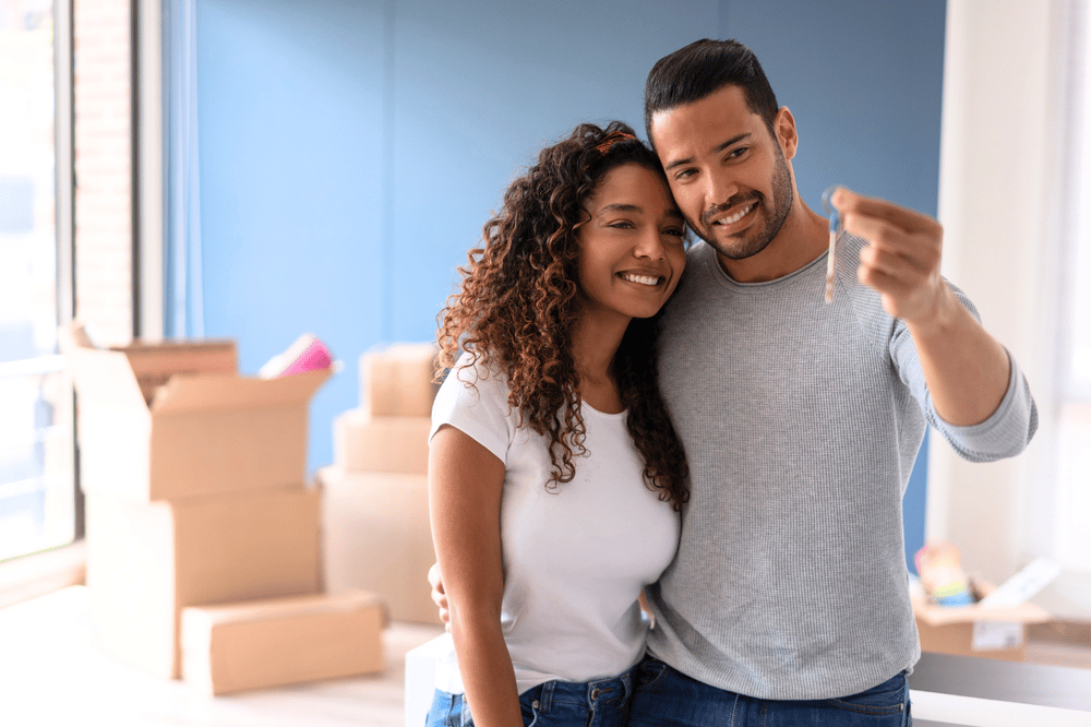 Smiling couple celebrates with a house key beside moving boxes.