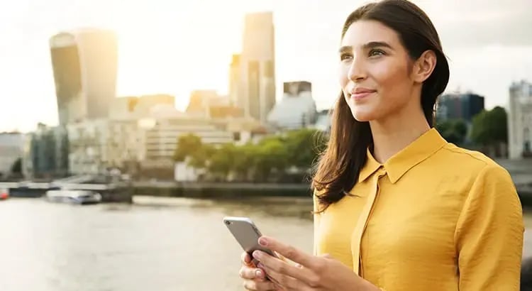 A woman standing outdoors with a cityscape in the background. She is holding a phone and smiling, indicating she is enjoying a pleasant moment in the city.