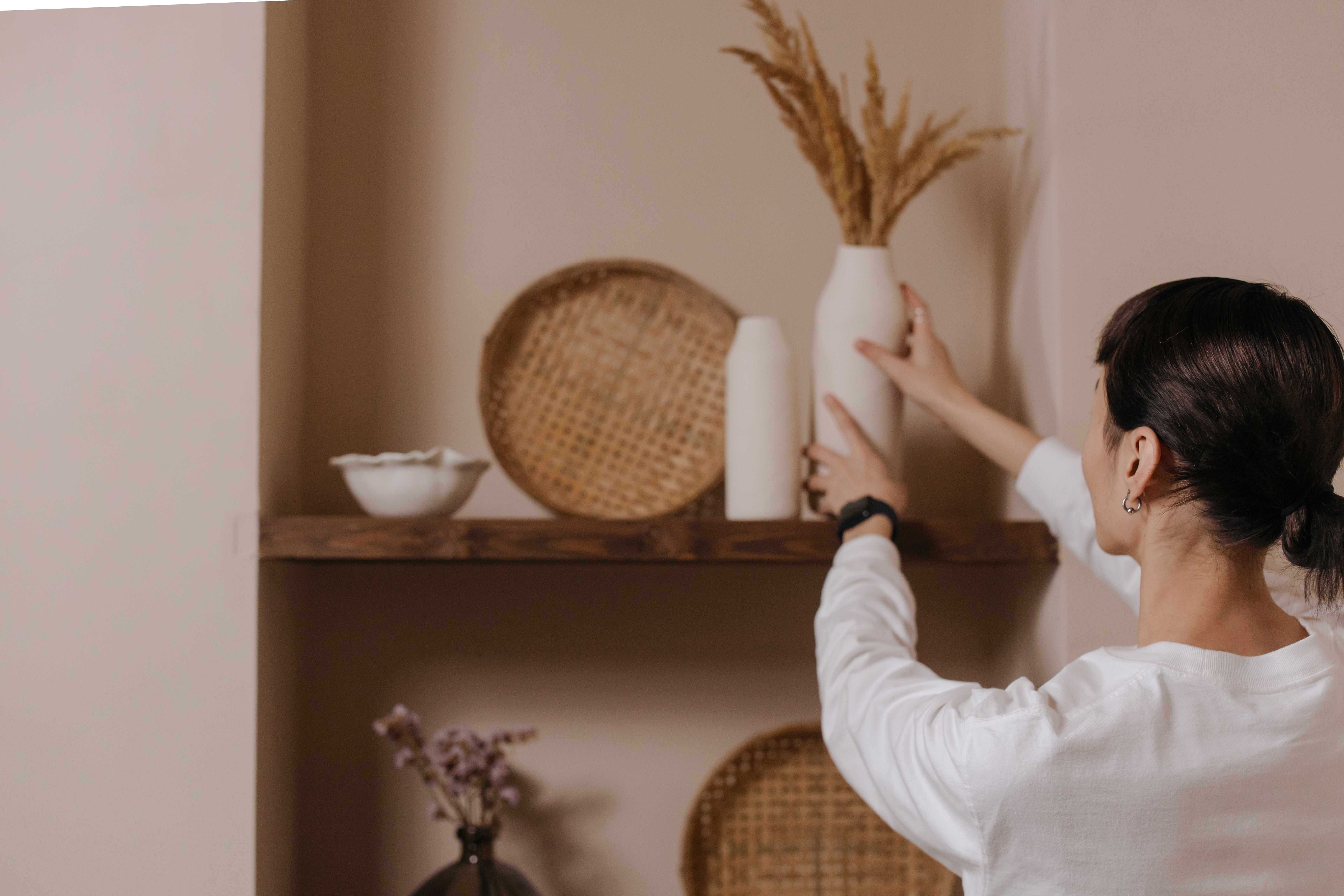 Woman styling a shelf with a white vase of dried grasses, woven baskets, and ceramic decor.