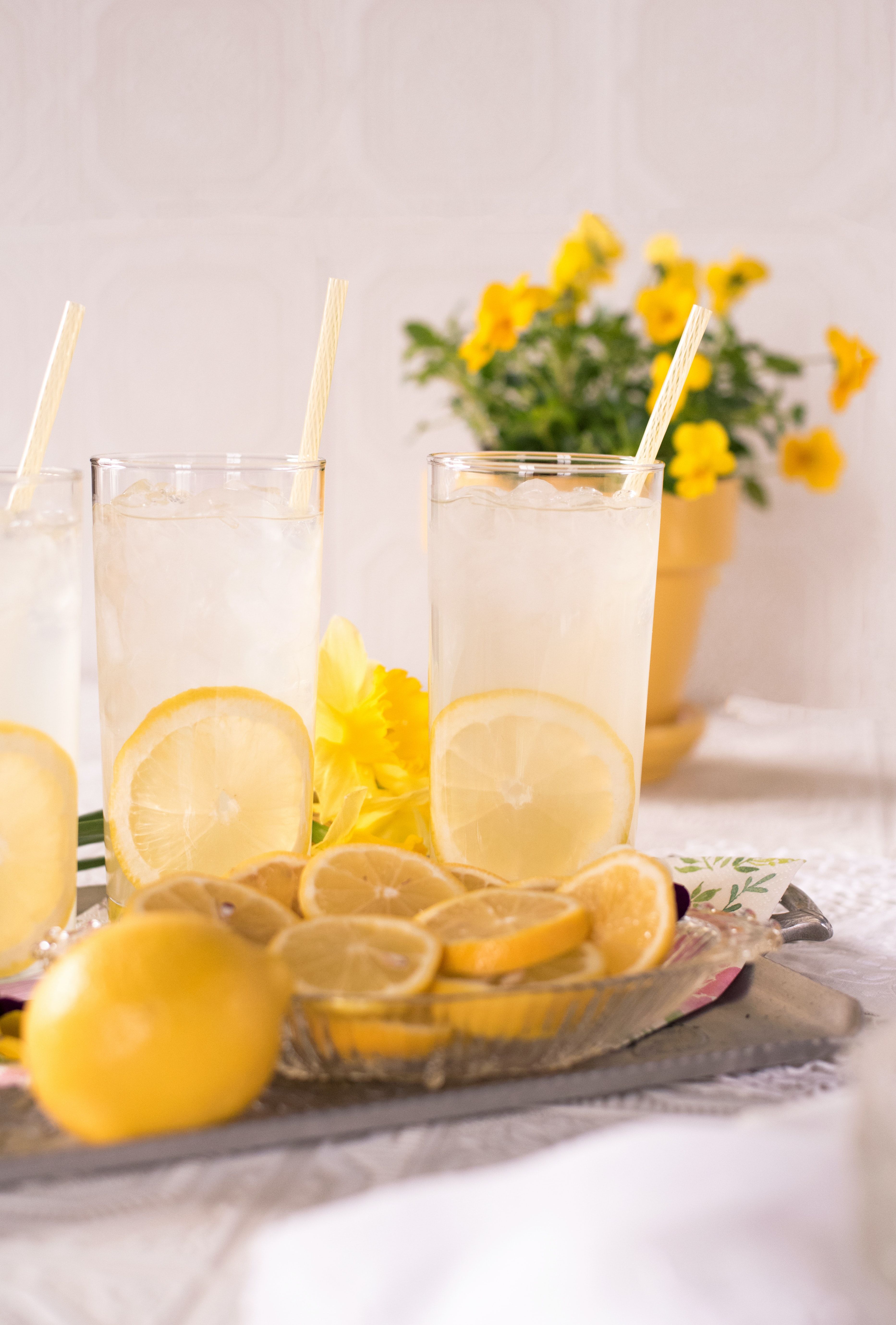 Iced lemonade with lemon slices beside a dish of fresh lemons and yellow flowers.