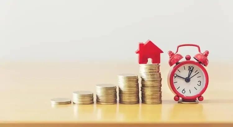 A stack of coins next to a small red house model and a red alarm clock, symbolizing financial planning or investment in real estate.