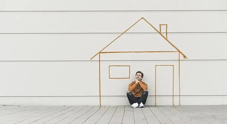 A person sitting against a wall with a drawing of a house in orange lines behind them. The drawing includes a roof, windows, and a door, suggesting a creative representation of home.