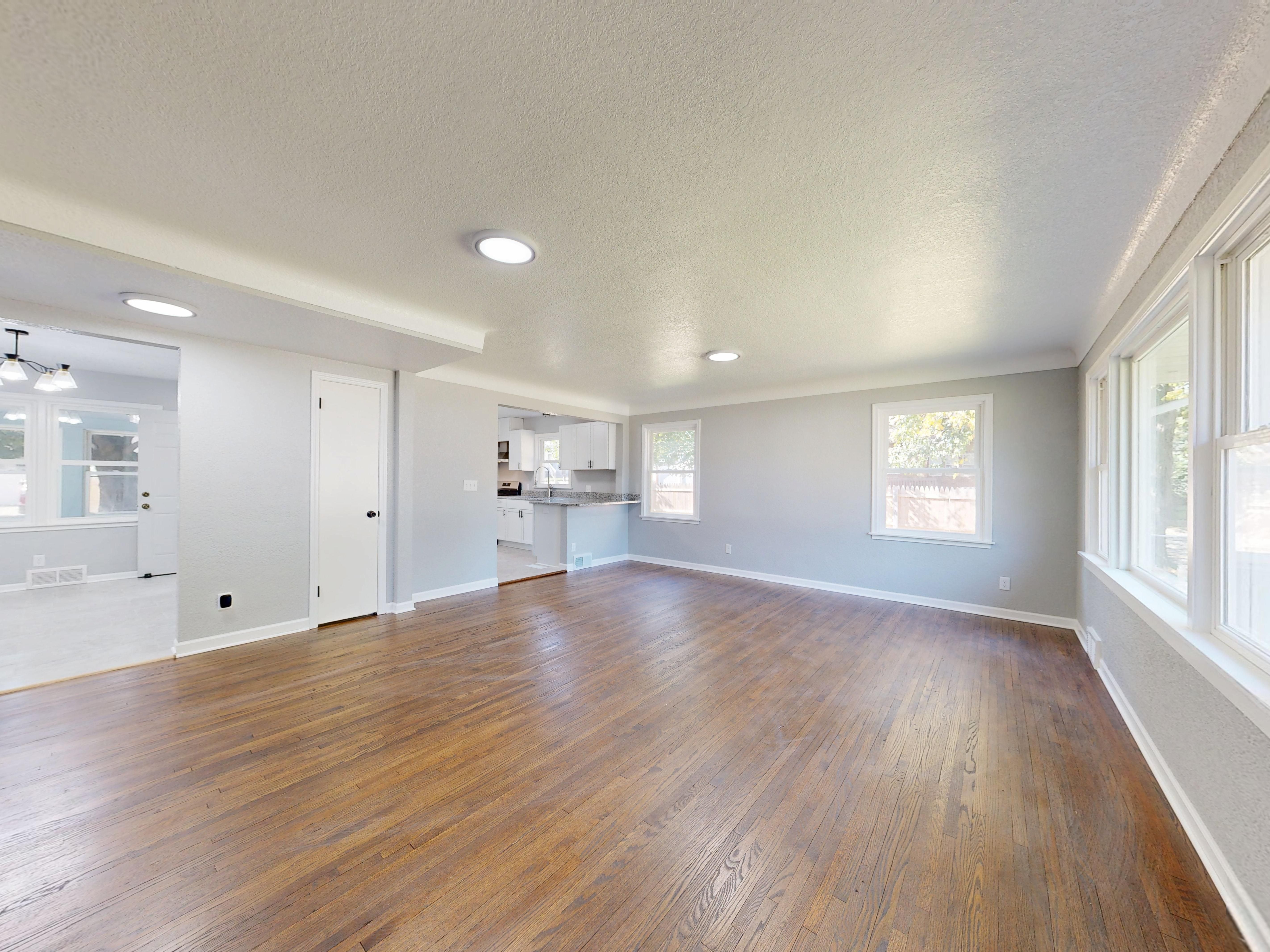Empty living room with hardwood floors, large windows, and an open view into the kitchen.