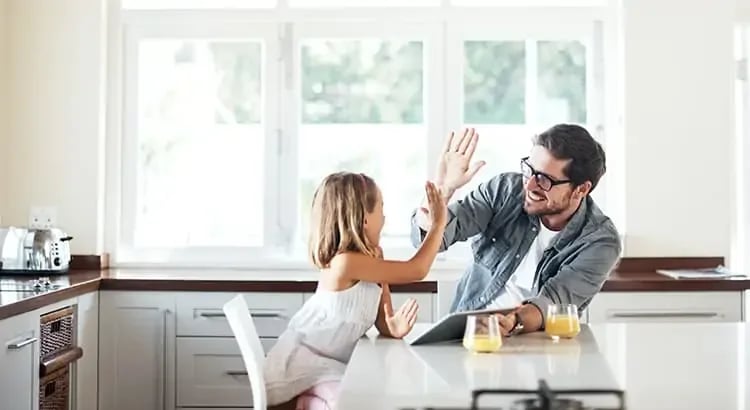 A man with his child sitting at a kitchen table, sharing a light-hearted moment. The child is raising their hand, possibly to high-five the father.