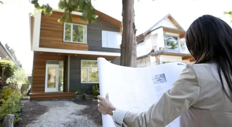  A person holding blueprints or plans while looking at a house under construction or renovation. The house has a modern design with large windows and wooden exterior elements.