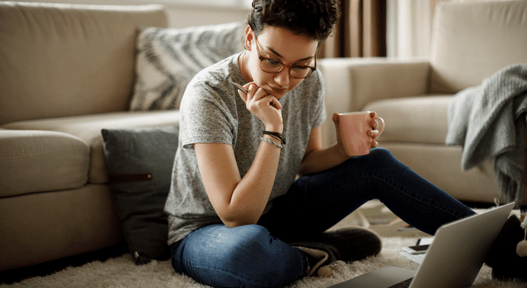 Person sitting on floor of beige living room with coffee and pen reading laptop