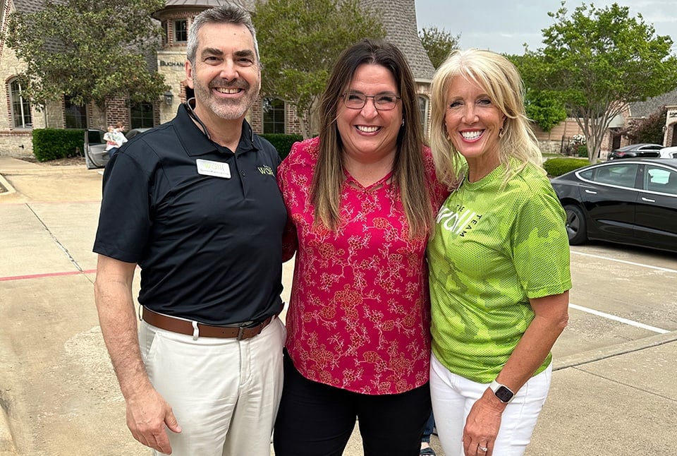 A man with white hair, a blonde woman, and a brunette woman with a house in the background