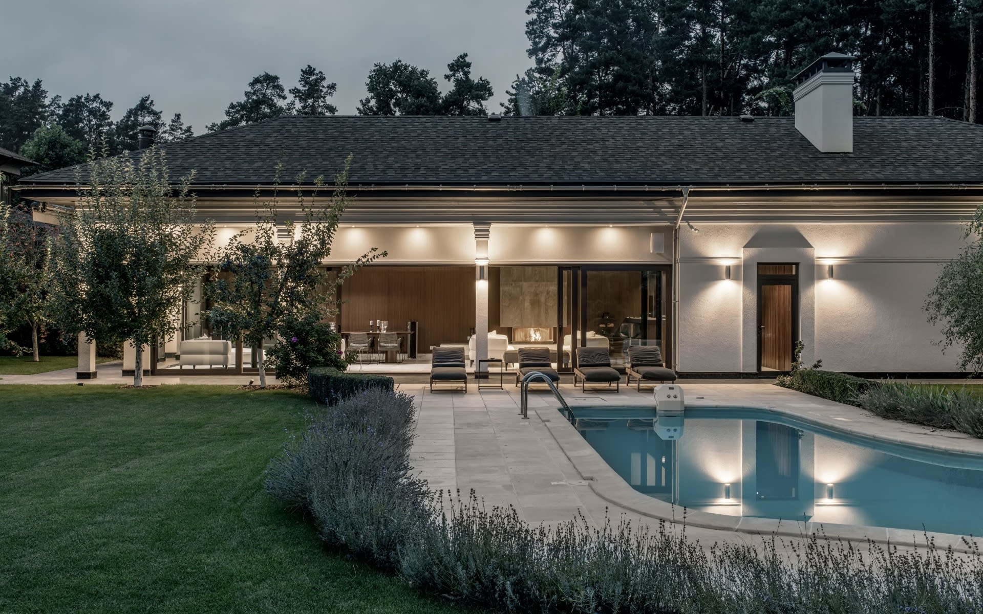 Modern home’s covered patio and illuminated rear facade beside a swimming pool at dusk.