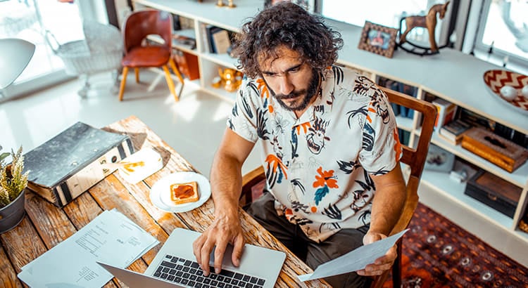 A man working at a desk with a laptop, notebook, and various office supplies. The setting is casual, and the man appears to be focused on his work.