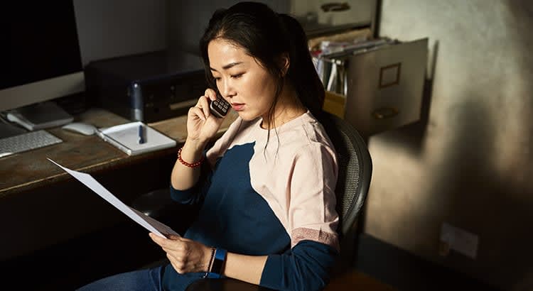 A woman, possibly working from home, sitting at a desk while talking on the phone and holding papers.