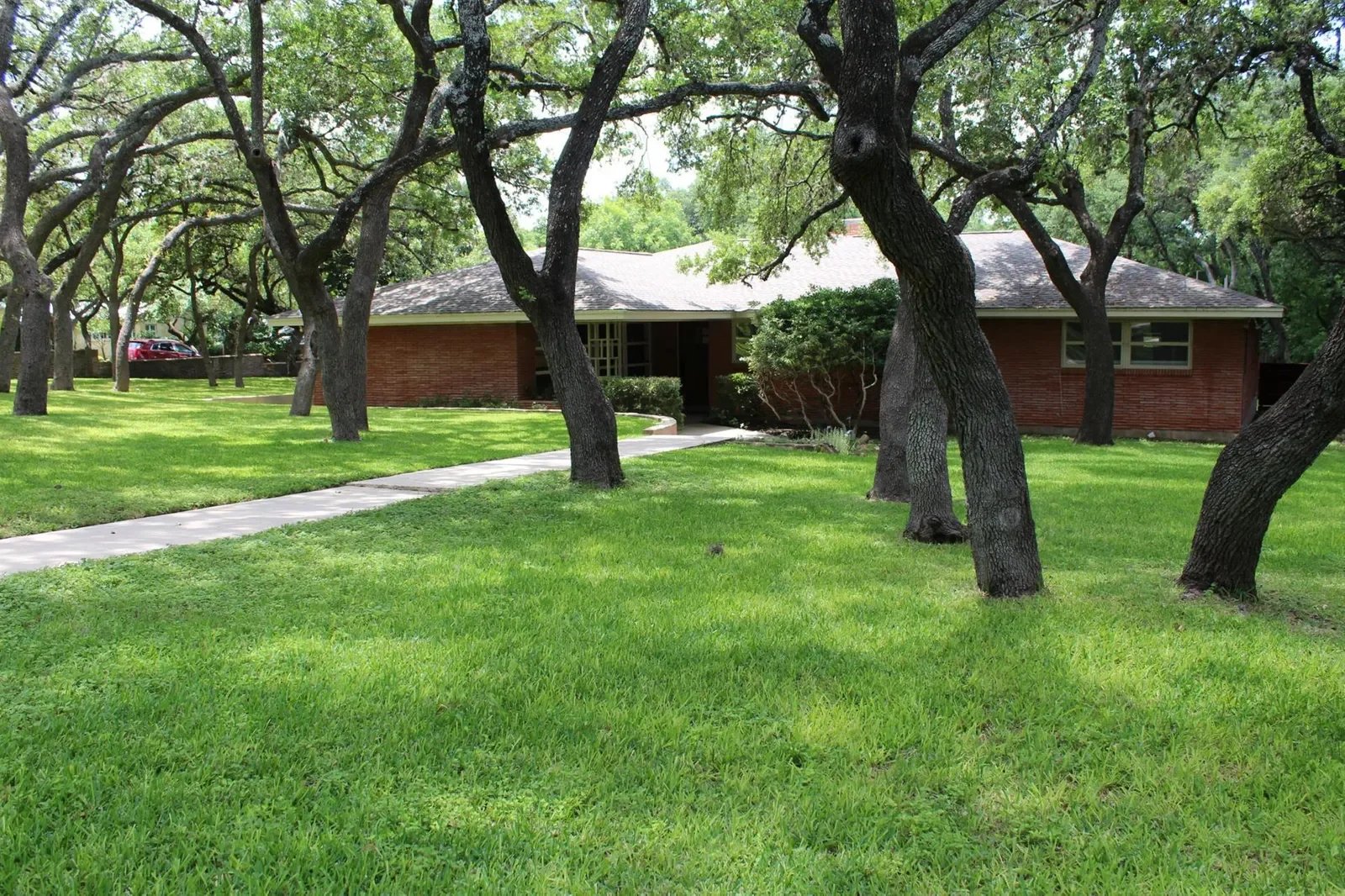 Single-story red brick house framed by mature oak trees, with a walkway across the lawn.