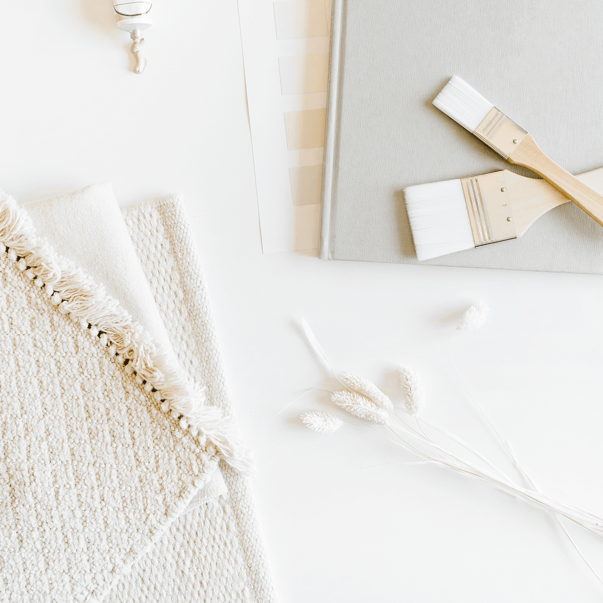 Paintbrushes and pale paint swatches beside textured cream fabric and dried grasses.