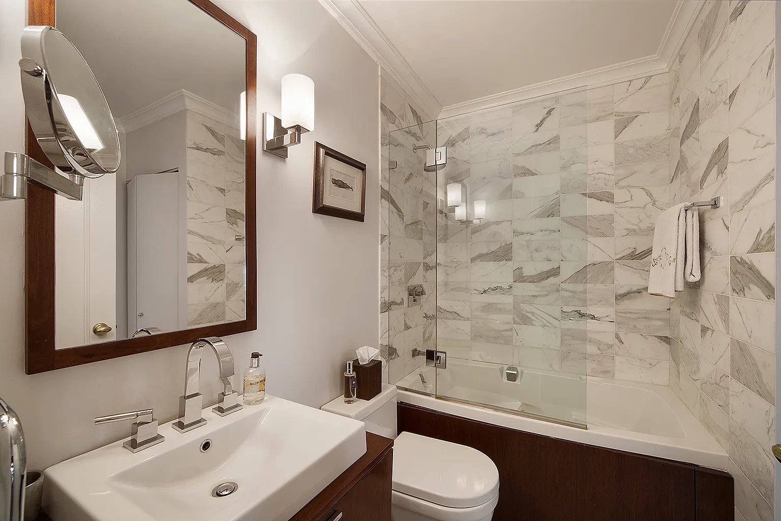 Bathroom with patterned marble tile, a glass-enclosed tub, and dark wood vanity.