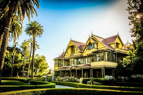 Yellow Victorian mansion with steep gables and a wraparound porch, framed by palm trees and manicured lawns.