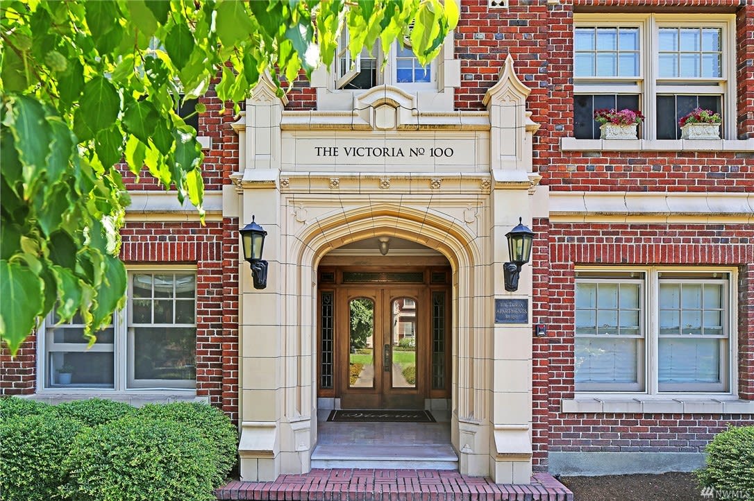 An ornate entryway of The Victoria, a luxury condo building, is framed by lush foliage and brickwork charm.