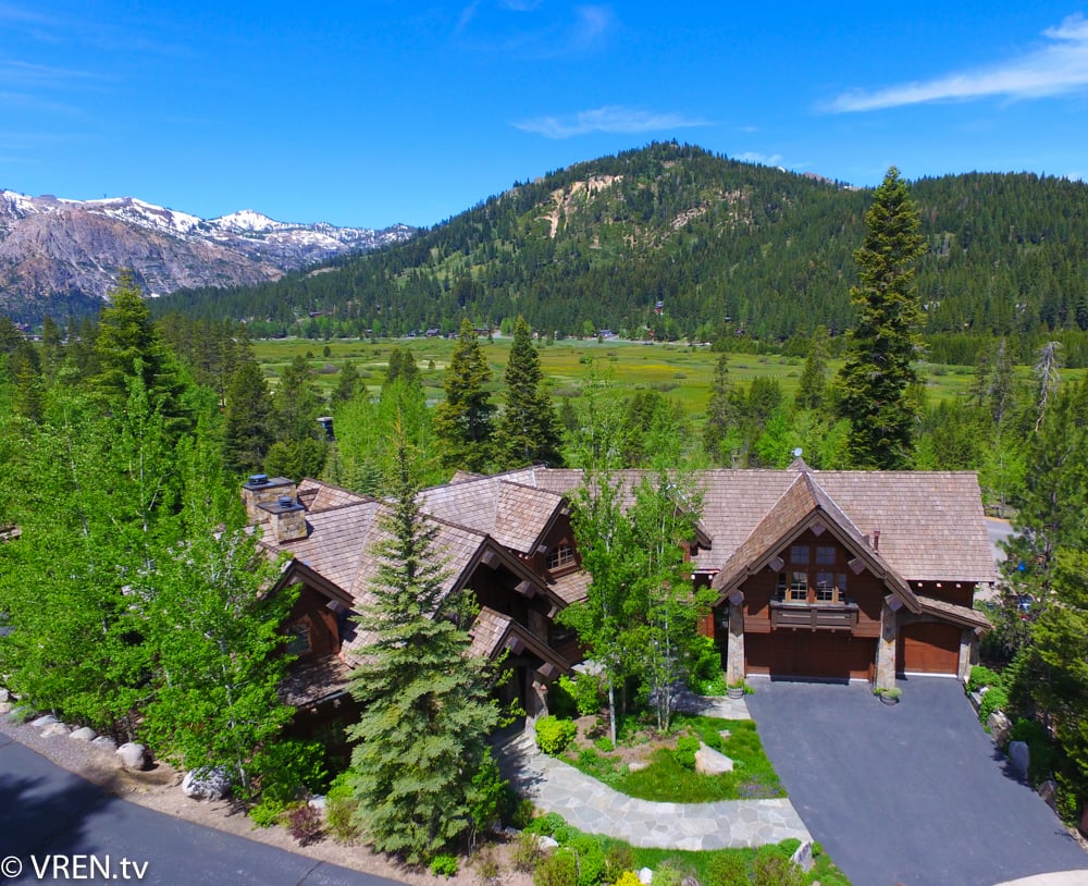 Aerial view of a gabled mountain home surrounded by trees, with a meadow and forested peaks beyond.