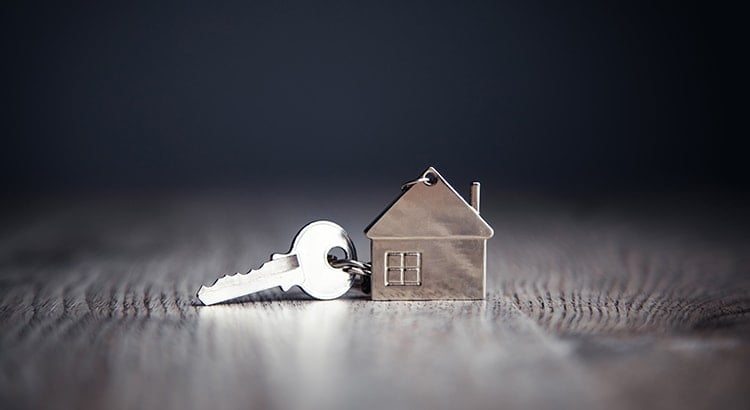 A house with a large key in the foreground. The background is dark and foggy, creating a dramatic effect. This image likely represents the concept of home ownership.