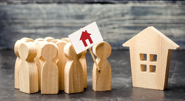 A wooden figurines representing people are grouped together, with one holding a sign featuring a red house icon. To the right of the group is a small, wooden house model with windows cut out.